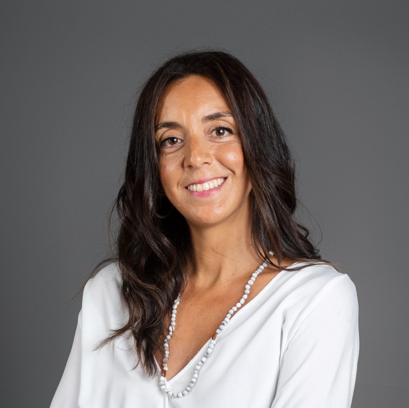 Retrato profesional de Laura Rivas sonriendo con camisa blanca sobre fondo gris.